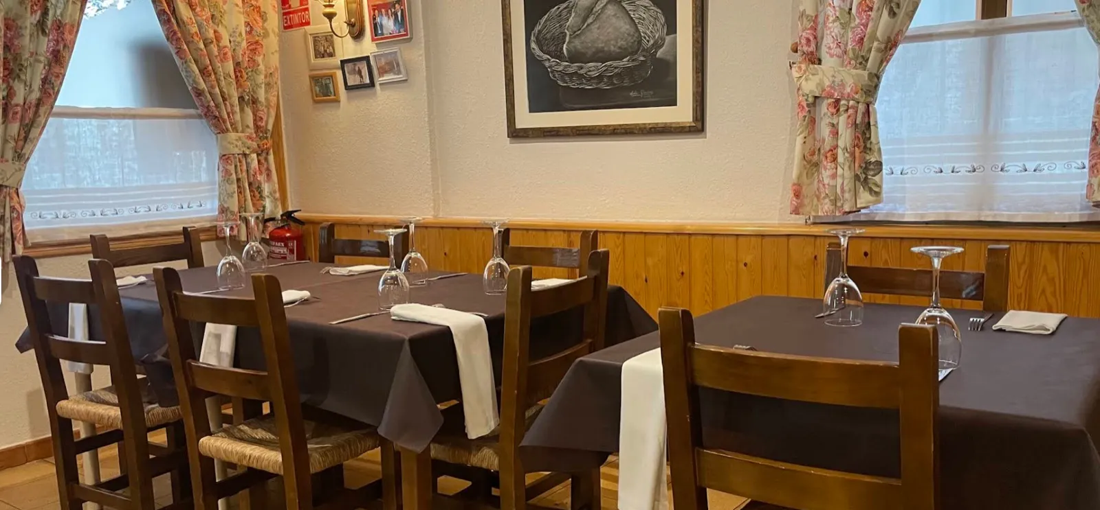 Comedor del Asador Ixarso en Benasque: mesas vestidas con mantel, sillas de madera, zócalo de madera y cortinas de flores, un ambiente acogedor de casa