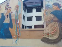Mural en la doble fachada de la plaza del Centro de Ciencias de Benasque: a la izquierda un mayordomo con flores en la cabeza y castañuelas de cintas de colores; a la derecha la dona de Benás bailando con el traje tradicional y el Aneto con su cruz al fondo