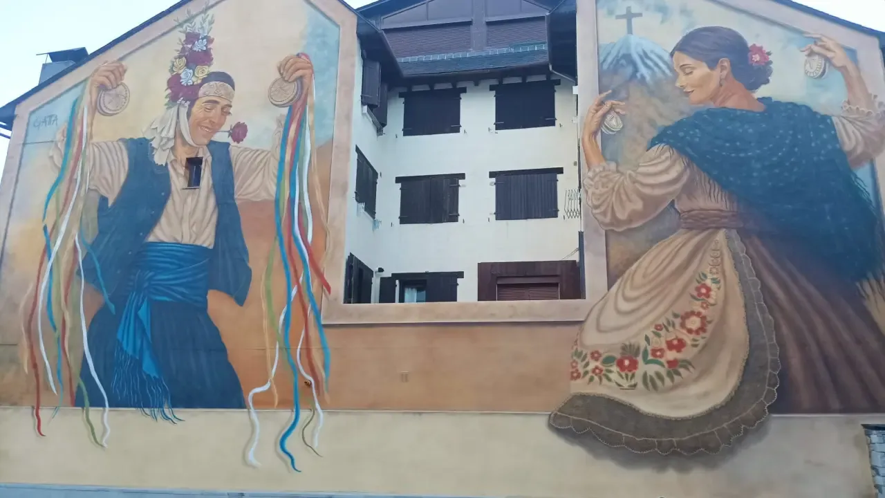 Mural en la doble fachada de la plaza del Centro de Ciencias de Benasque: a la izquierda un mayordomo con flores en la cabeza y castañuelas de cintas de colores; a la derecha la dona de Benás bailando con el traje tradicional y el Aneto con su cruz al fondo